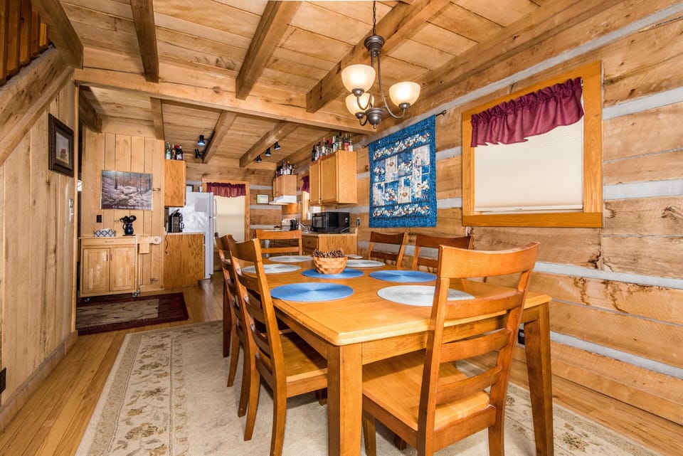 Dining area with a wooden table, six chairs, blue and beige placemats, and quilt wall hanging in log cabin.