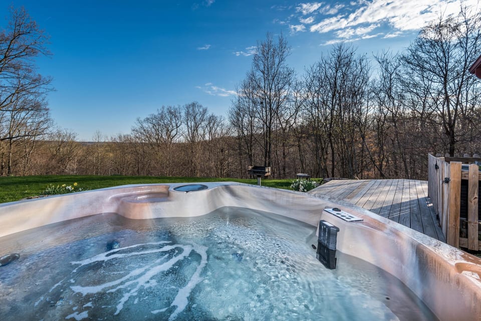 Outdoor hot tub bubbling beside a wooden deck, overlooking leafless trees under a blue sky.