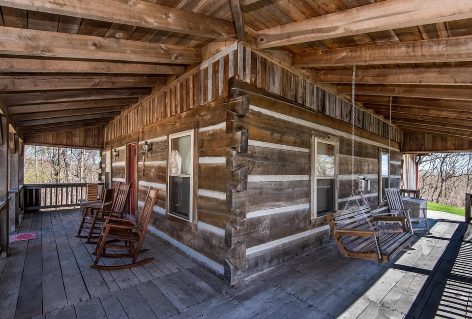 Wide wraparound porch with weathered wood planks, rocking chairs, and a wooden swing overlooking trees.