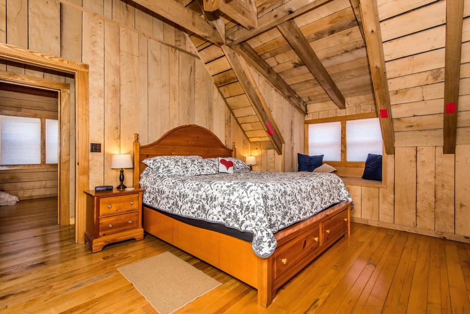 Upper bedroom with vaulted wood ceiling, angled beams, built-in window bench, and wooden bed with floral bedding.