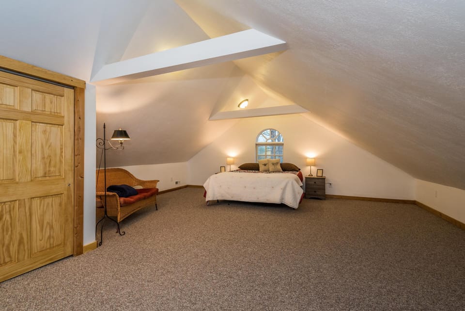 Attic bedroom under sloped ceilings with beige carpet, wooden closet door, wicker loveseat, and arched window.