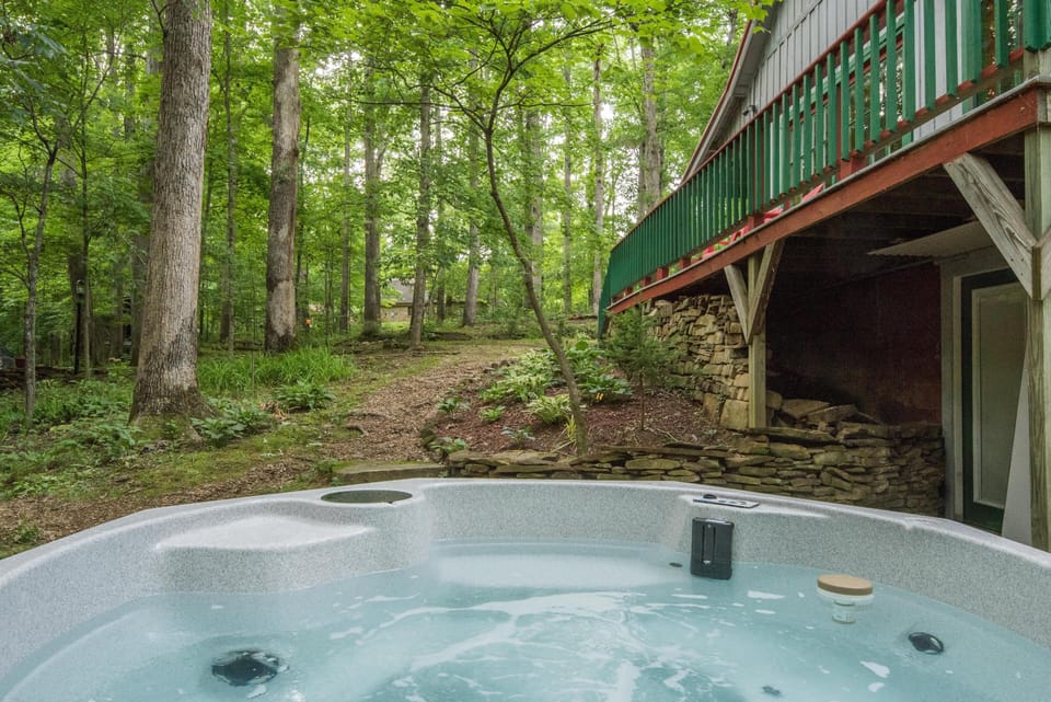 Bubbling hot tub sits beside a stone retaining wall under a green and red wood deck, surrounded by tall trees.