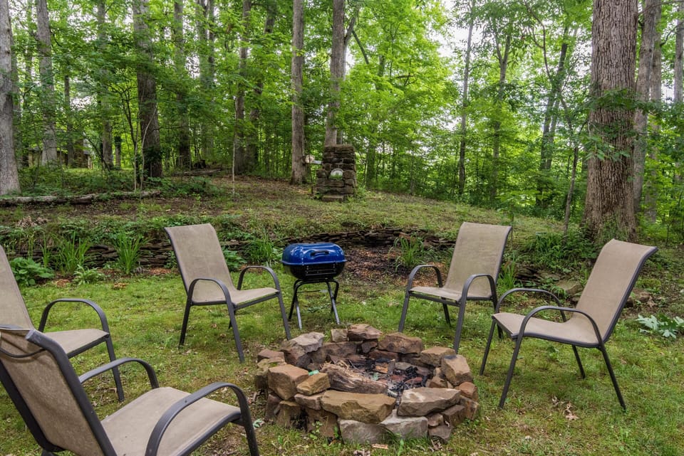 Stone firepit encircled by five beige mesh chairs on grassy clearing with tall trees in background.