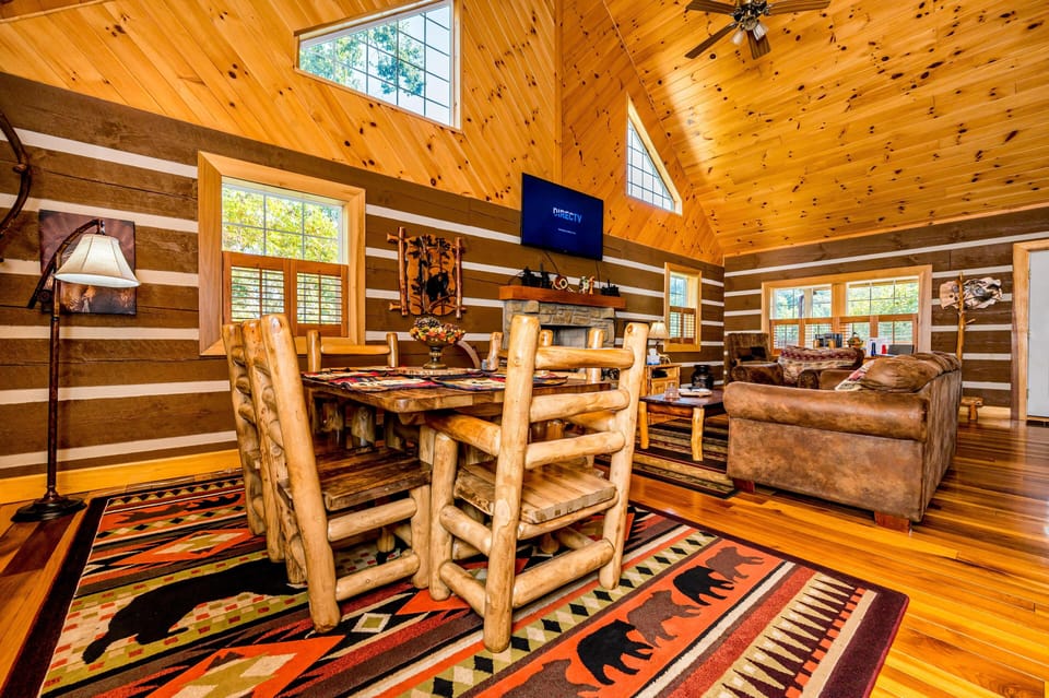 Dining area with log chairs and table set on a patterned rug, vaulted pine ceiling, and large windows.