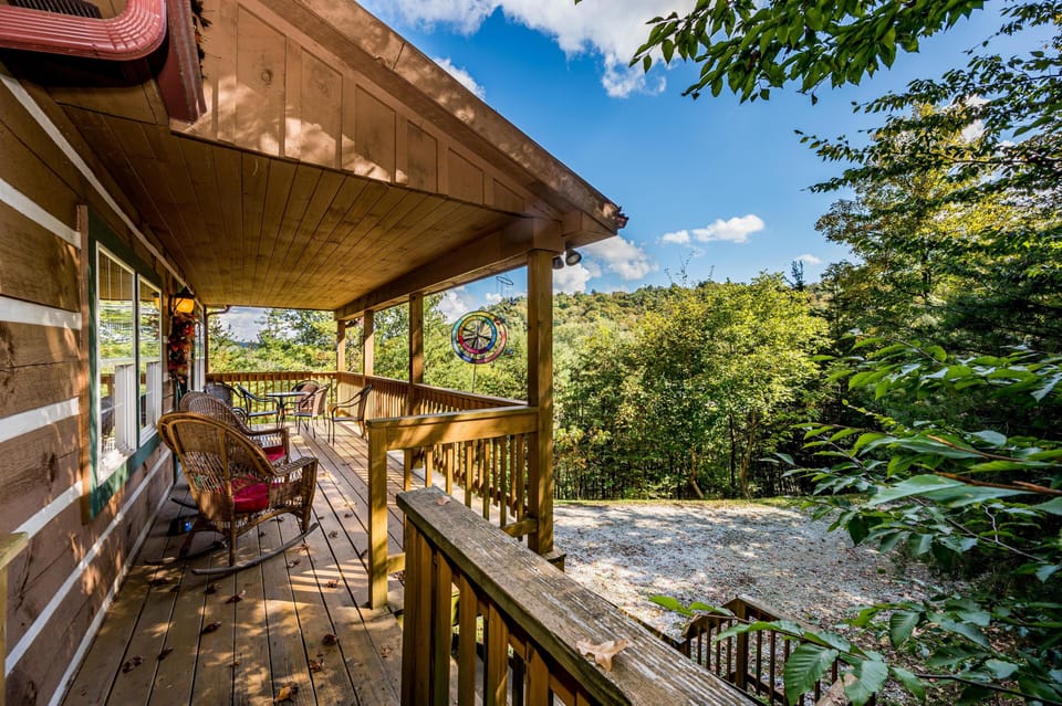 Covered wooden deck with wicker rocking chairs, metal bistro set, and forested hillside views.