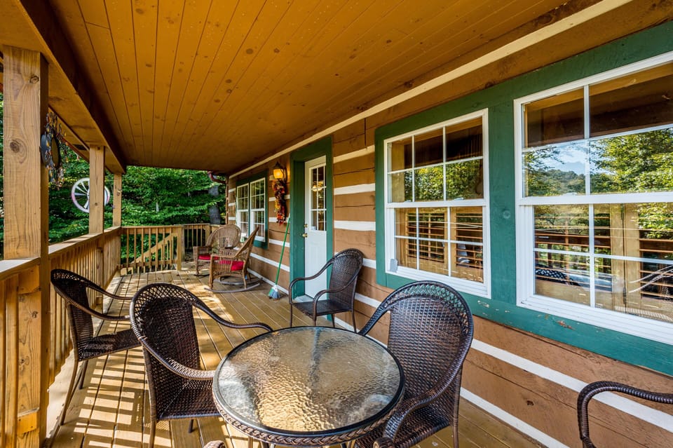 Long wooden porch with round glass table, four woven chairs, rocking chairs, and green-trimmed cabin windows.