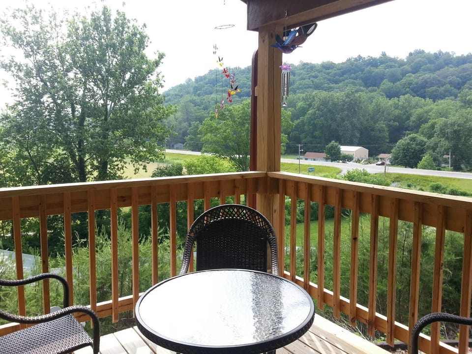 Wooden balcony with woven chairs and a round glass table overlooks green hills and a nearby highway.