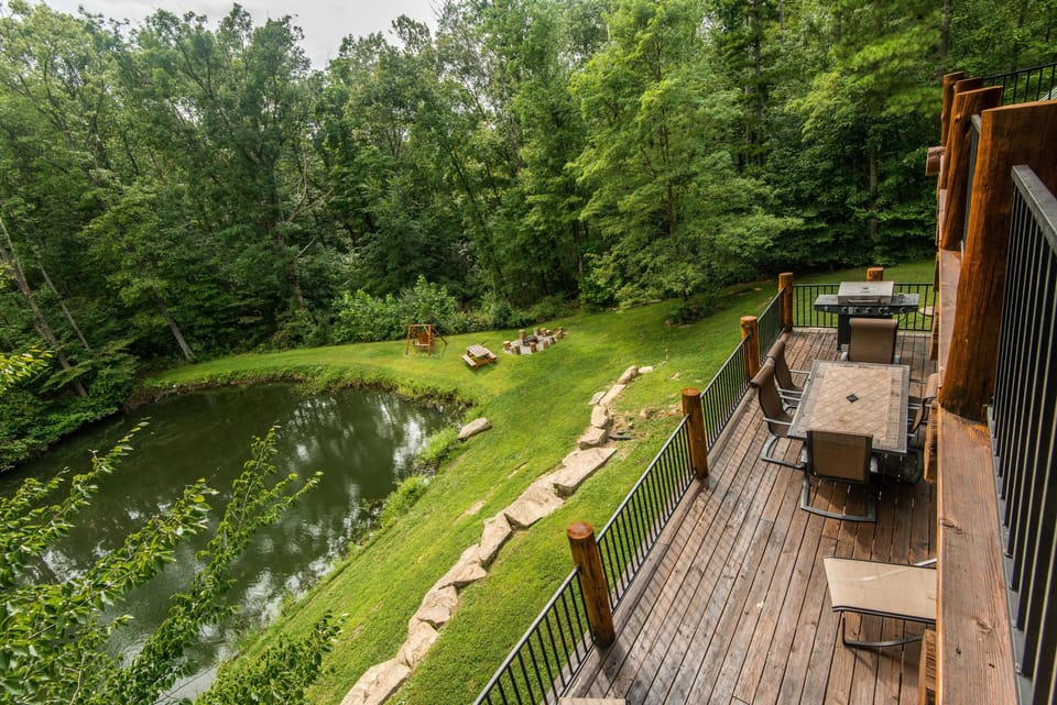 Upper wooden deck with metal railings, outdoor dining set, grill, and views of stocked pond and firepit area.