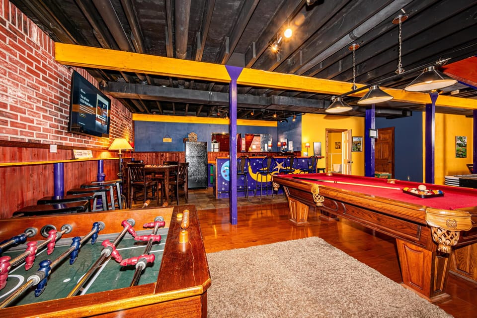 Game room with red-felt pool table, foosball, purple and yellow beams, brick wall, and a starry painted bar.
