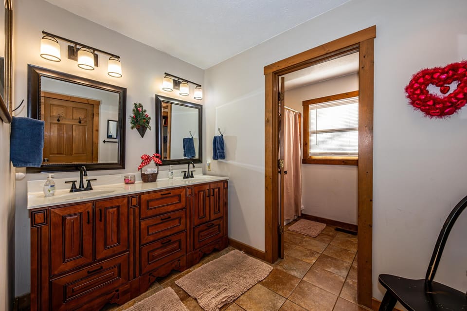 Bathroom with wood-trimmed mirrors, double sink vanity, tiled floor, and shower area with window.