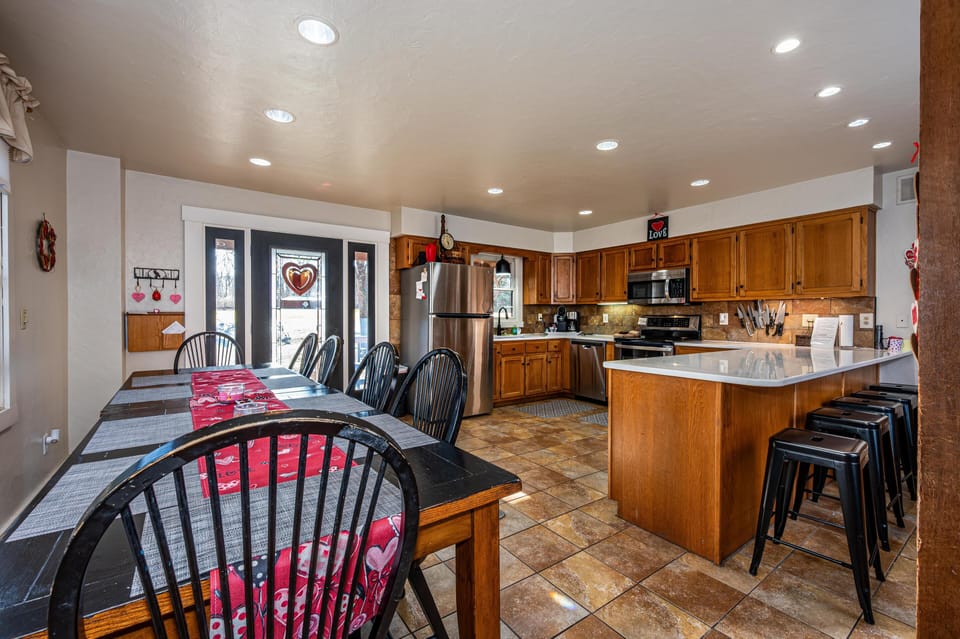 Dining area with black wooden chairs and a long table beside French doors, adjacent to a kitchen with wood cabinets and tiled floor.