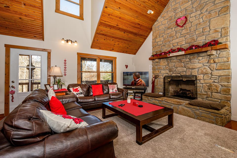 Vaulted pine ceiling and stone fireplace frame a living room with dark leather sofas and a dark wood coffee table.