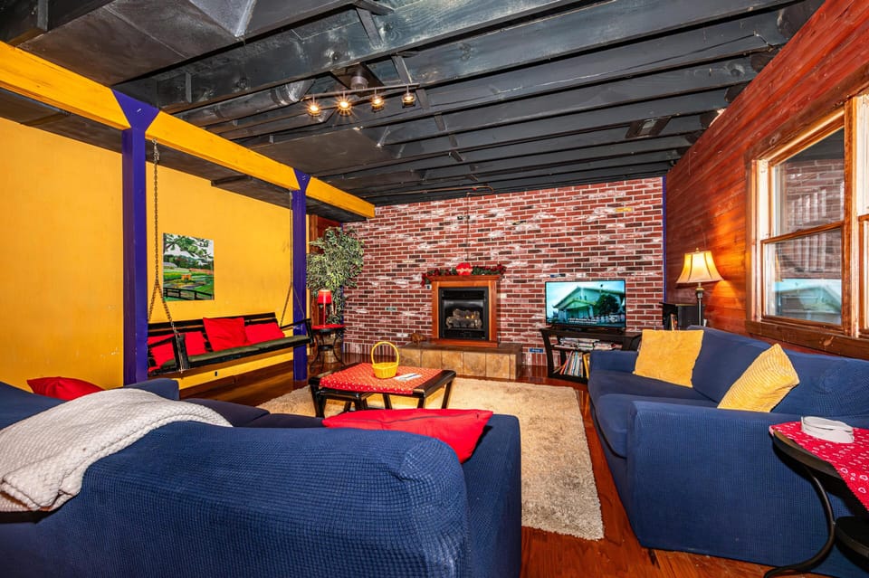 Living room with exposed black ceiling beams, a brick fireplace, blue sofas, and a red and yellow swing bench.