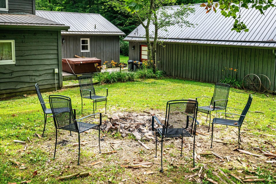 Metal mesh chairs encircle a stone firepit on patchy grass between gray wood-paneled buildings and orange daylilies.