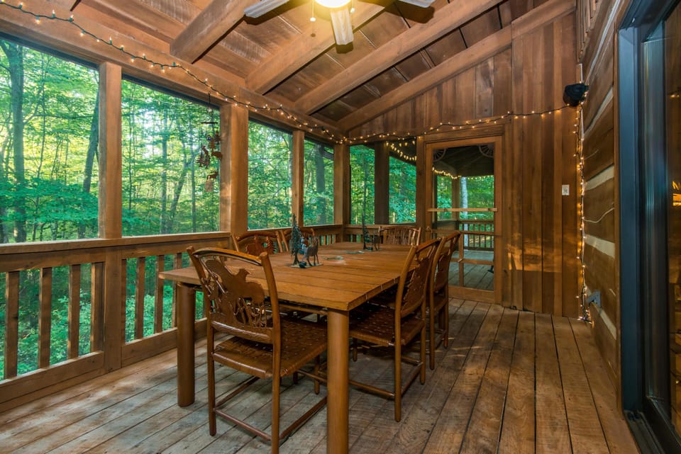 Wooden dining table and rustic chairs fill the screened porch with string lights and forest views beyond.