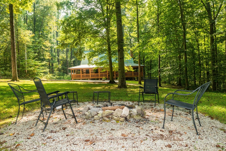 Outdoor fire pit area with black metal chairs encircling stones, set on white gravel and surrounded by trees.