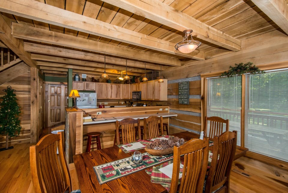 Dining area with wooden table, pine furniture, patterned runner, and large window with blinds overlooking porch.