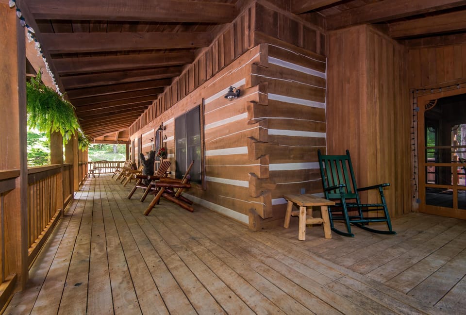 Wide wooden porch with log walls, exposed beams, green rocking chair, hanging ferns, and multiple Adirondack chairs.