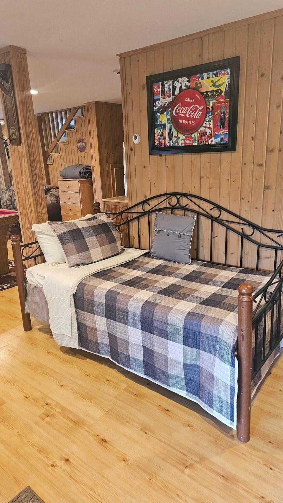 Daybed with black metal frame and plaid quilt sits against pine wood-paneled wall beneath colorful Coca-Cola artwork.