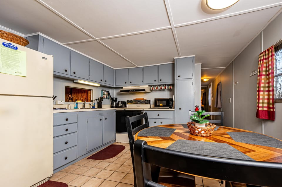 Dining table with black chairs sits beside a kitchen featuring pale blue cabinets, beige tile floor, and white appliances.