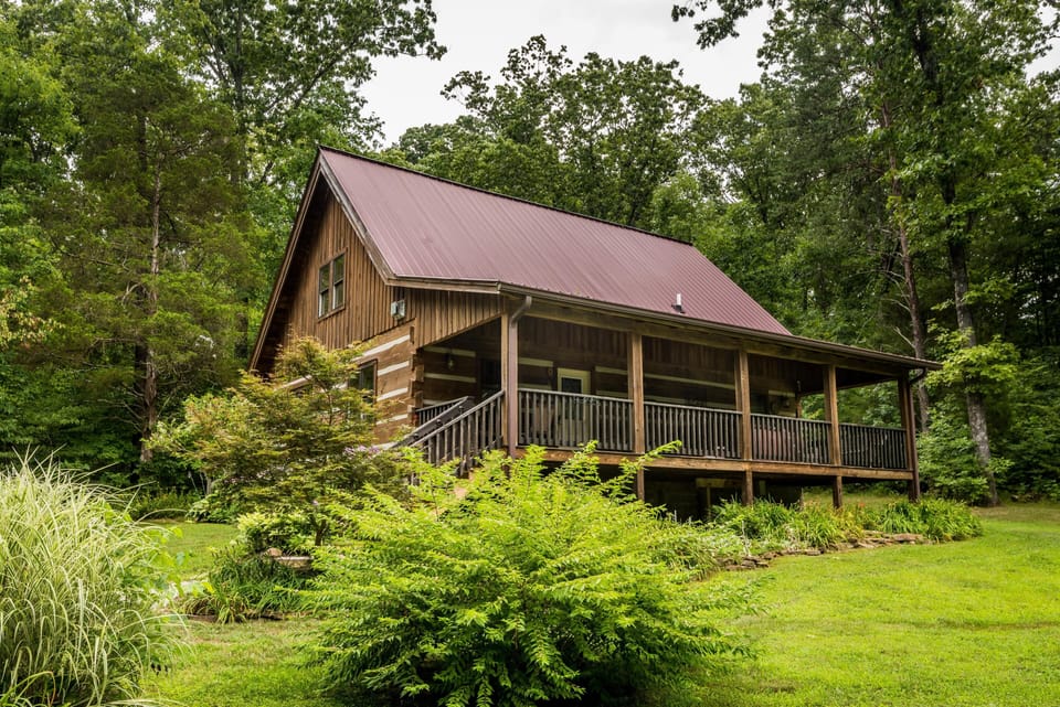Side view of the log cabin with a red metal roof, covered porch, wooden railing, and lush green landscaping.