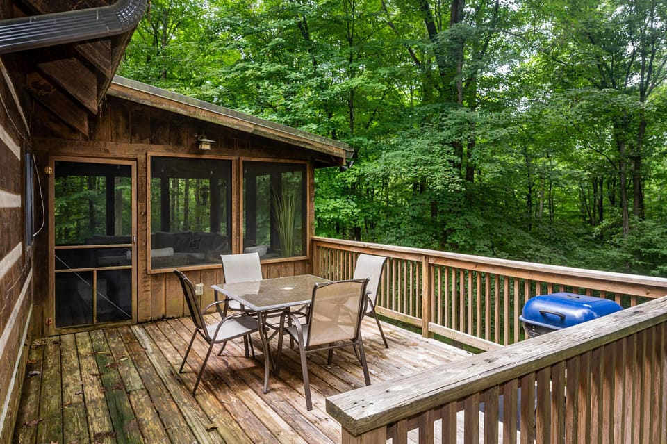 Wood deck with glass-top dining table, four mesh chairs, blue charcoal grill, and forest views.