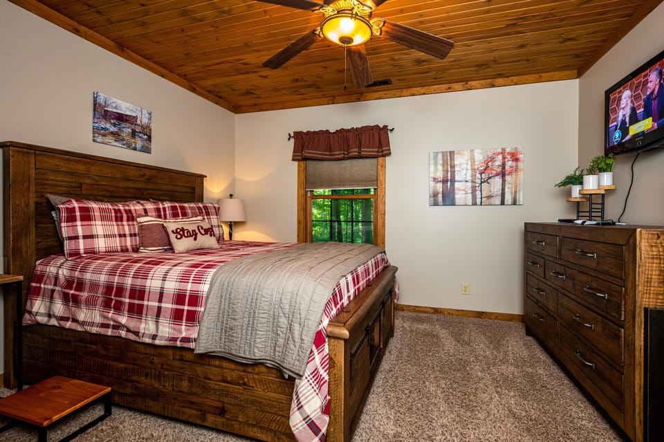Master bedroom with wooden bed frame, red plaid bedding, ceiling fan, and flat-screen TV above rustic dresser.