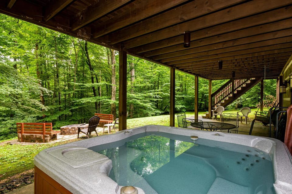 Covered patio with hot tub, metal and plastic chairs, and brick fire pit framed by dense green forest.
