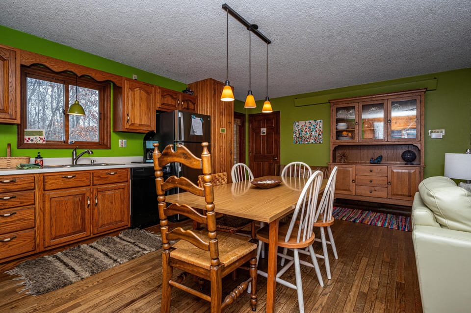 Dining area with a wooden table, mix of ladder-back and spindle chairs, hanging amber pendant lights, and green walls.