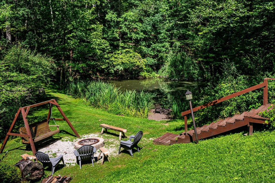 Gravel fire pit area with Adirondack chairs, wooden swing, rustic bench, and pond dock surrounded by trees.
