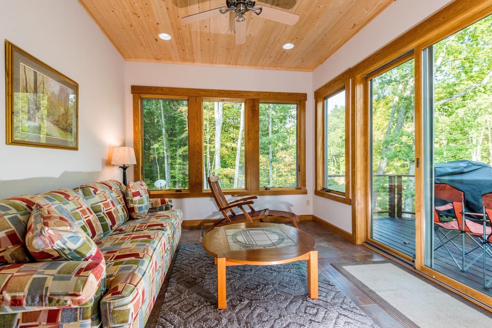 Sunroom with patterned sofa, wood-framed windows, ceramic tile flooring, and sliding door to deck with grill.