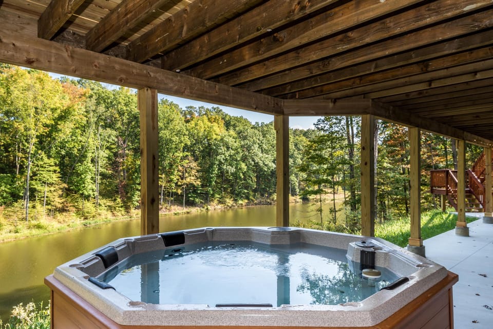 Outdoor hot tub with wood framing sits under covered deck overlooking a calm lake and dense green trees.