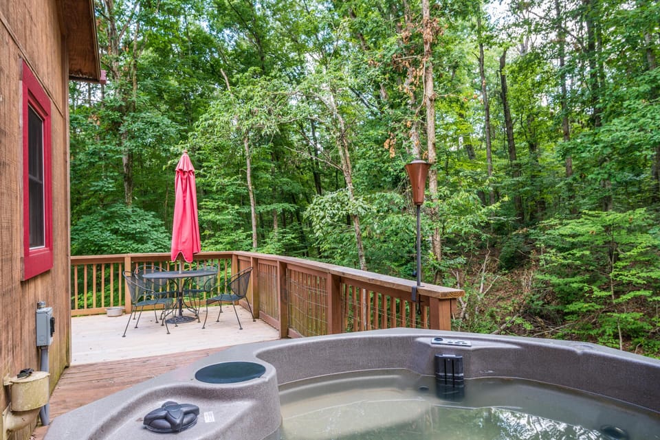 Wood deck with a filled gray hot tub, black metal table and chairs, red umbrella, and forested backdrop.
