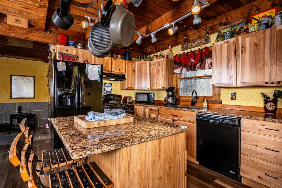 Granite-topped island with wooden stools faces black appliances and knotty pine cabinets in the kitchen.