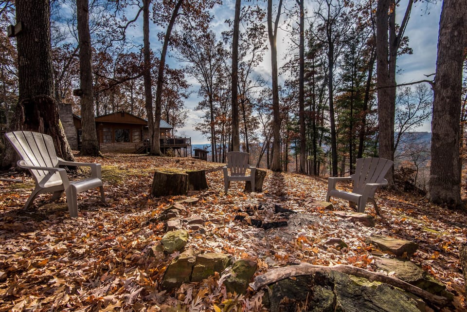 Leaf-covered fire pit area with three plastic Adirondack chairs and a view of the cabin among tall trees.