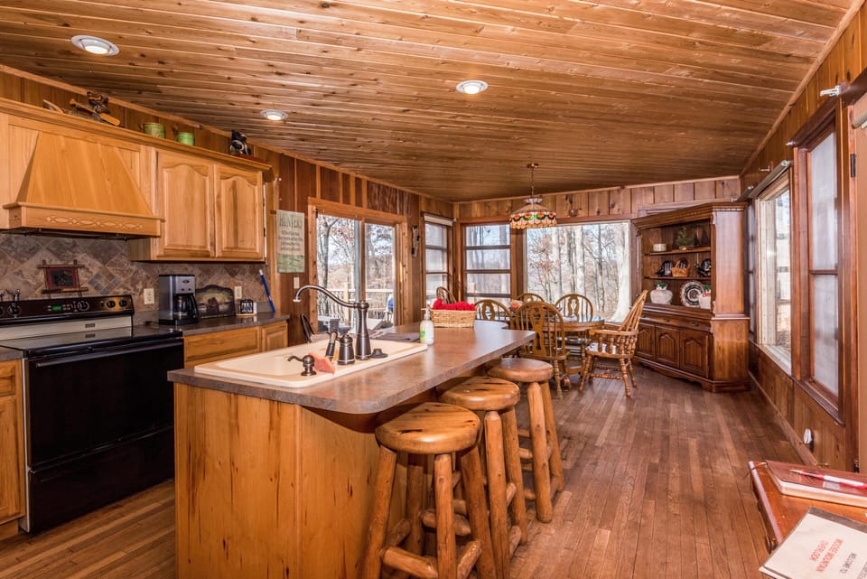 Kitchen island with rustic wooden stools and a double sink faces the dining area and forest views through windows.