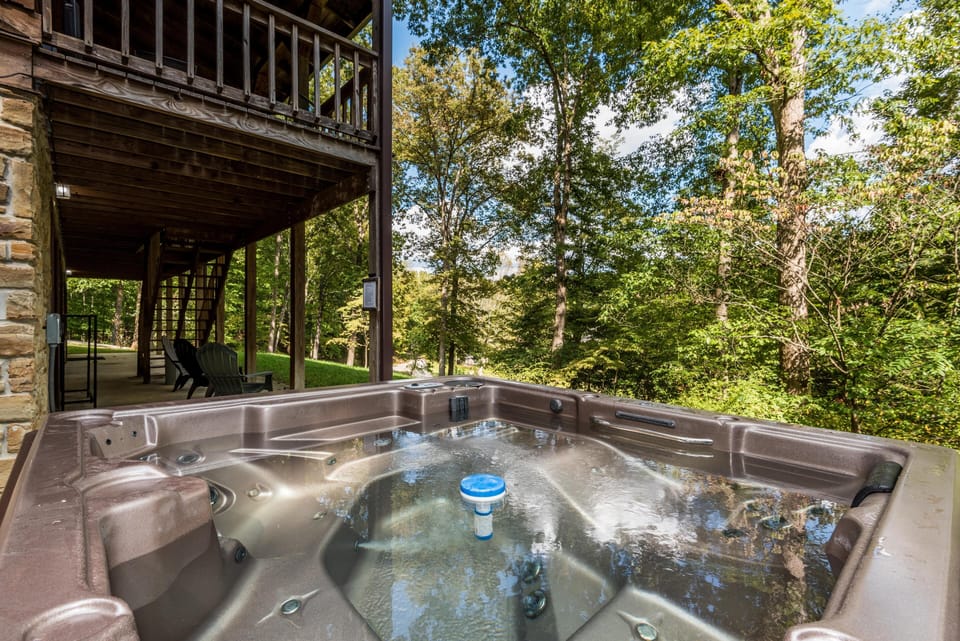 Outdoor hot tub with built-in seating beneath a wooden balcony, surrounded by lush green trees.
