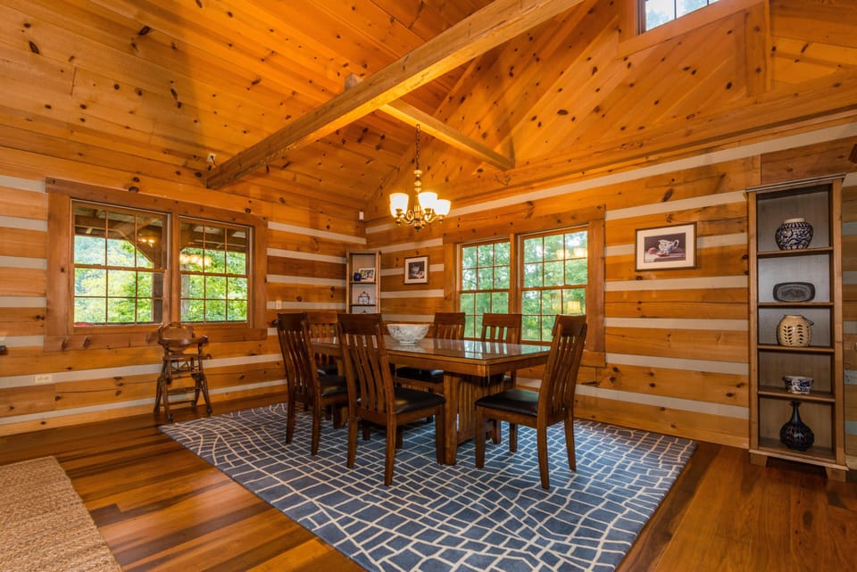 Wood-paneled dining room with vaulted ceiling, rectangular wooden table, eight chairs, and patterned blue rug.