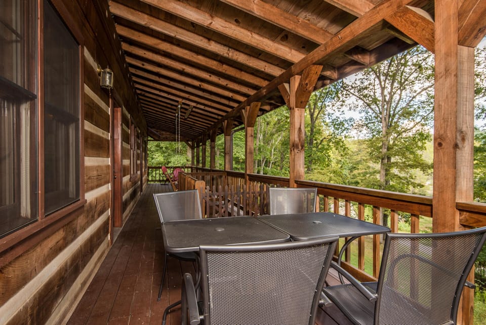 Covered wooden deck with metal dining table, mesh chairs, Adirondack chairs, and views of leafy trees.
