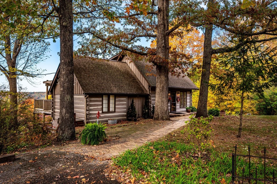Brick pathway winds past mature trees leading to a log cabin with large windows and seasonal wreaths by the entrance.