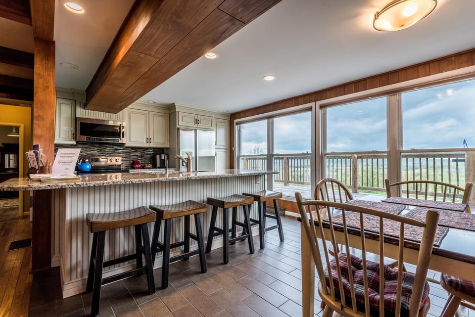 Kitchen breakfast bar with four wooden stools, granite countertop, and large windows overlooking the deck and hills.