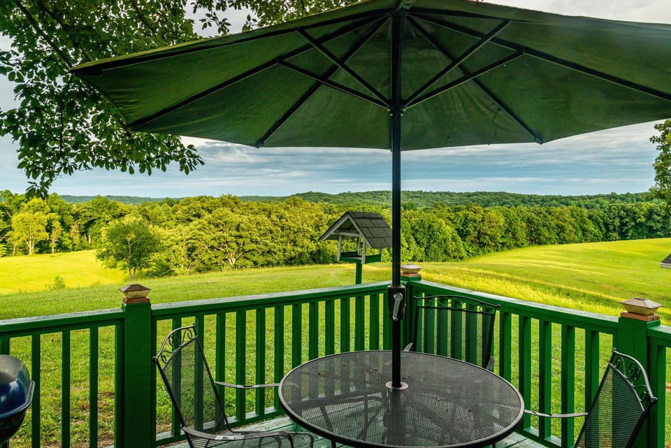Poolside seating with black metal table and chairs shaded by a large green umbrella, overlooking rolling green fields.