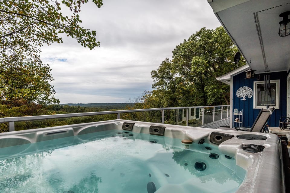 Outdoor hot tub bubbling beside a blue exterior wall, overlooking wooded hills and glass railing on the deck.