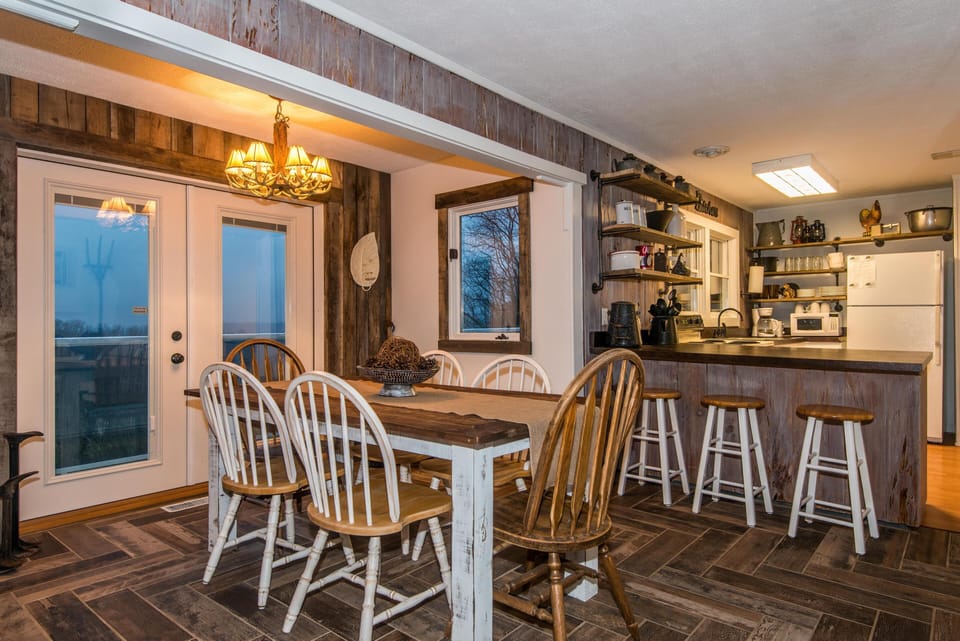 Dining area with mixed wood chairs and a farmhouse table, adjacent to a kitchen with open shelving and wood-paneled island.