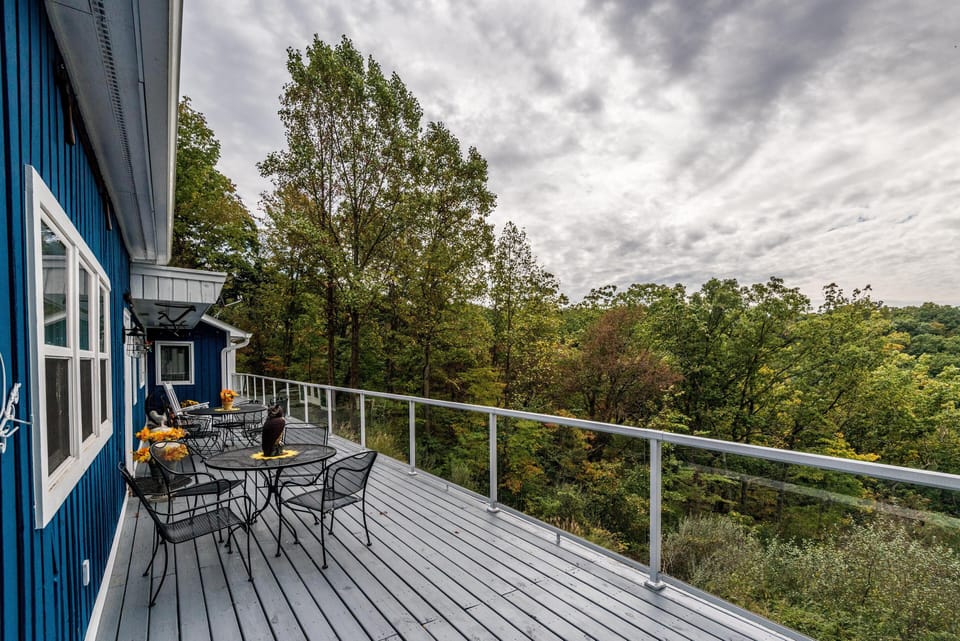 Blue exterior wall with white trim lines the spacious deck, outfitted with black metal tables and chairs beside a glass railing.