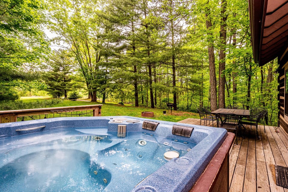 Outdoor deck with a bubbling blue hot tub, black metal dining set, and lush green trees beyond the railing.