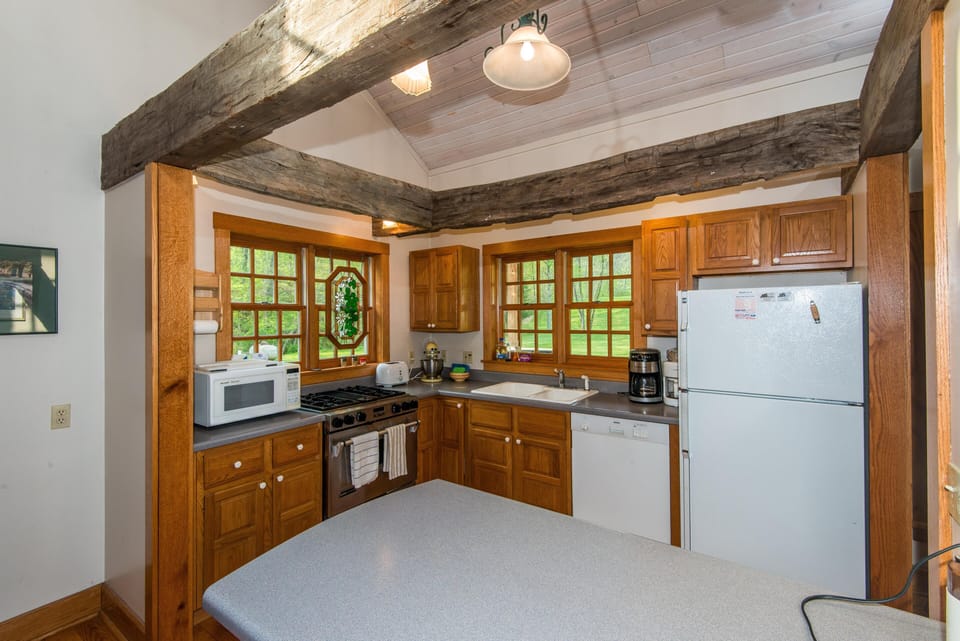 Kitchen with oak cabinets, gray countertops, two wood-framed windows, and exposed rustic ceiling beams.