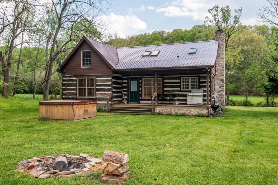 Backyard view of the log cabin with a covered porch, stone chimney, hot tub, charcoal grill, and fire pit on green lawn.