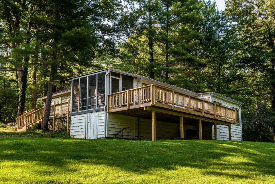 Elevated wooden deck with railings overlooks a green lawn and is adjacent to a screened porch surrounded by trees.