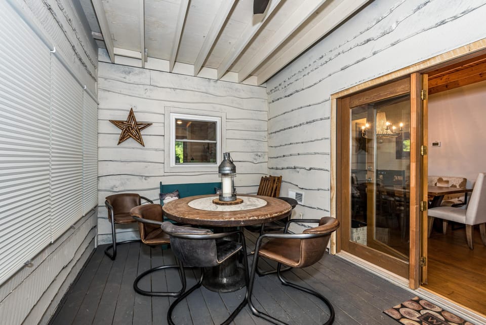 Screened porch with painted log walls, round wooden table, leather armchairs, and a lantern centerpiece.